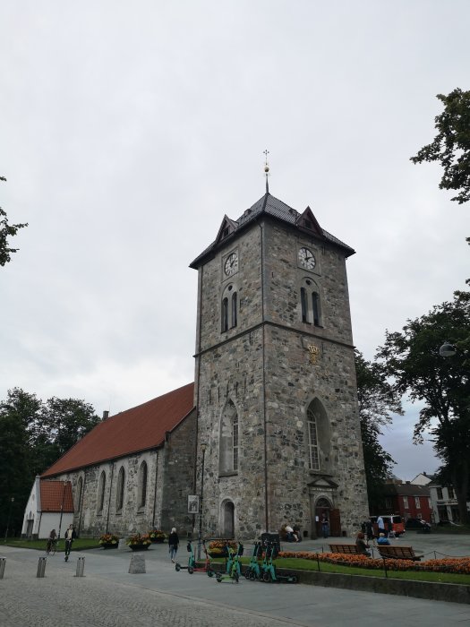 Kyrka i Trondheim med stentorn, klocka och spetsigt tak, omgiven av blomsterarrangemang och parkerade elsparkcyklar.