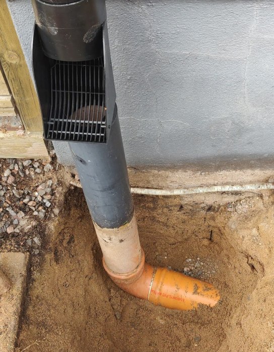 Grävd jord vid husgrund där ett stuprör möter en orange dräneringsrör i fuktig sand.