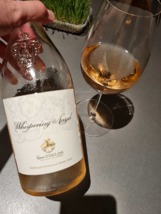 Flaska rosévin märkt "Whispering Angel" och ett glas med rosévin på en mörk yta, med gräs i bakgrunden.