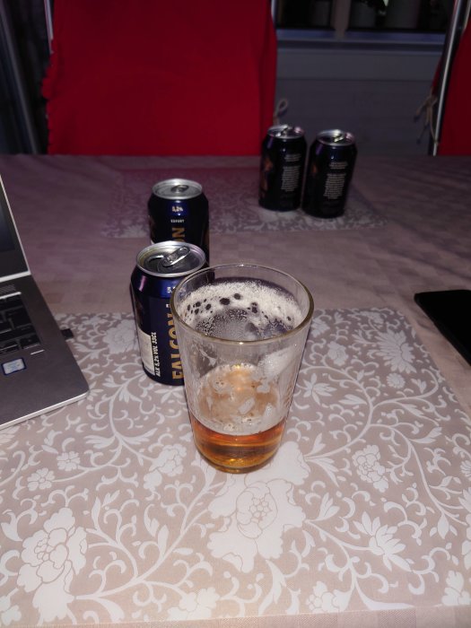 Halvt tömt ölglas med Falcon-öl framför en laptop, omgiven av tre oöppnade Falcon-ölburkar på ett mönstrat bord.