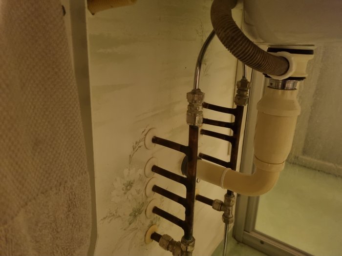 Röranslutningar till handdukstork med synlig vattenlås och tillförselrör vid vägg med tapet.