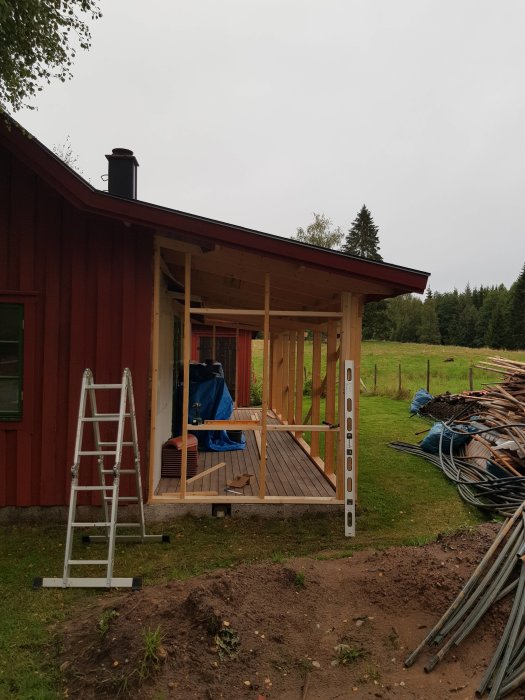 Nyreglad gavelvägg och veranda på ett rött hus med takskorsten, stege och byggmaterial synligt.