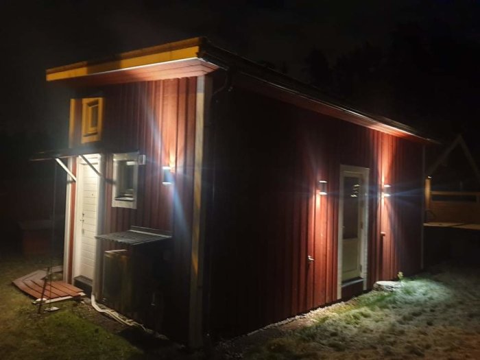 Nyligen installerad utomhusbelysning på en röd stuga med vit dörr under kvällstid.