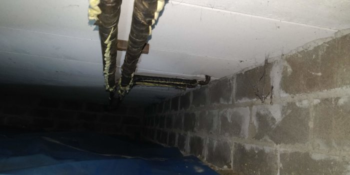 Isolerade rör som går längs en källarmur och tak, synlig fukt på murverket.