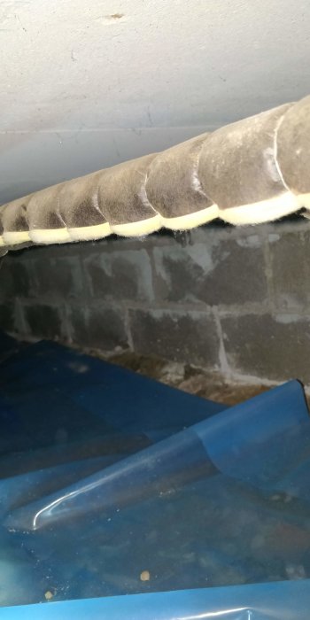 Isolerad ventilationsrörning mot en mörklagd tegelvägg över plastskyddad yta.