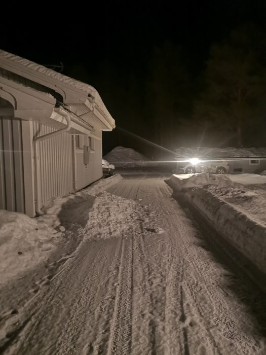 Belyst snöig uppfart vid hus med bil och träd i bakgrunden på natten.