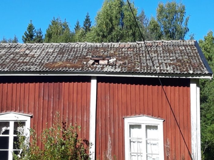 Äldre rött trähus med skadat tak av tegelstenar där några saknas, omgivet av grönska under solig himmel.