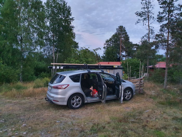 Bil parkerad utomhus med limträbalkar på takräcket och en person synlig inne i bilen.