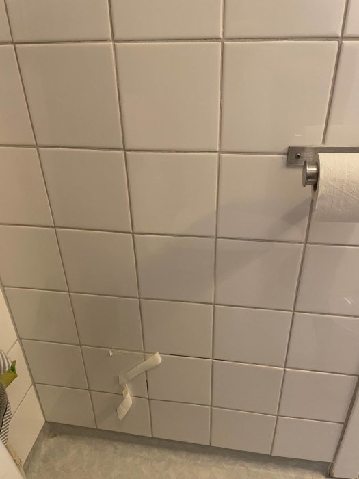 Vitt kakel i badrum med toalettpappershållare och rulle där en bit hänger ner.