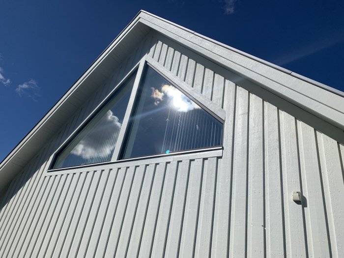 Vit husvägg med stort trekantigt fönster mot en klarblå himmel.