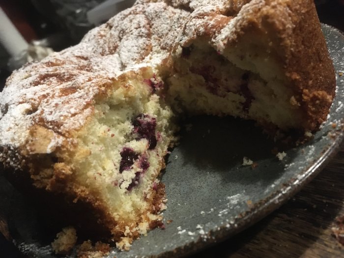 Hembakad blåbärskaka med synlig ihopsjunkning i mitten, pudrad med florsocker, på mörk bakgrund.