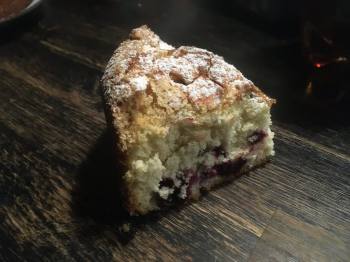 En portion blåbärskaka med synliga blåbär och strösocker ovanpå, mot en mörk bakgrund.