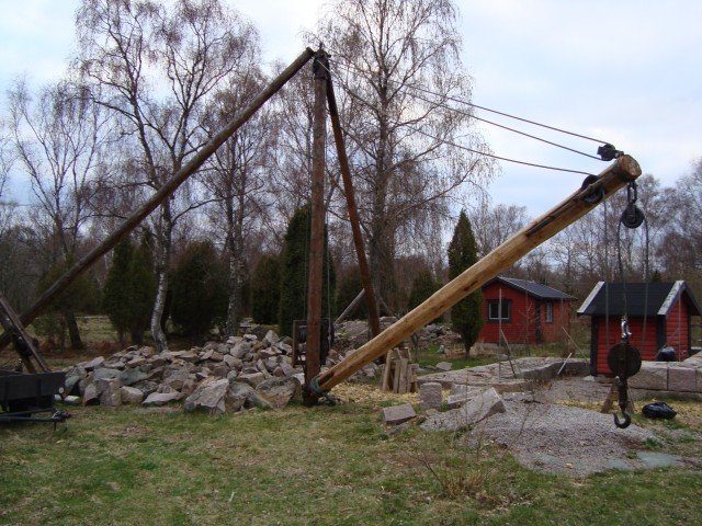 En hemmagjord kran konstruerad av trä och rep vid en stenhög i en trädgård.