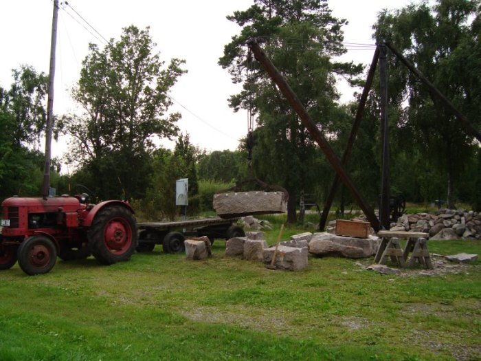 En gammal röd traktor med en släpvagn som lyfter en stor sten vid en byggplats med stenar och träd i bakgrunden.