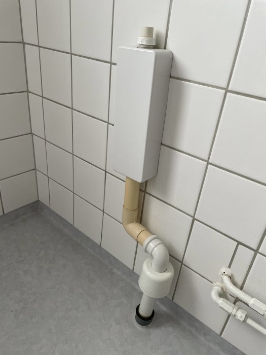 Väggmonterad avloppsstam och rör i badrum med vita kakelväggar och grått golv.