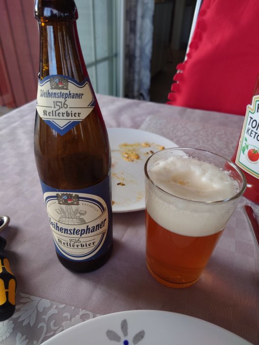 Weihenstephaner ölflaska och ett glas öl på ett bord bredvid en tom tallrik med smulor.