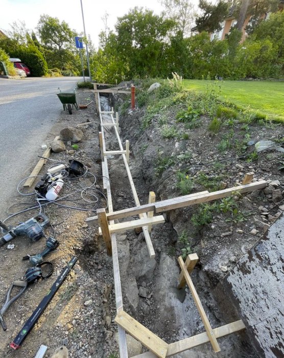 Utgrävning och uppställda träformar för en stödmur vid sidan av en altan, med armeringsjärn i berg och byggverktyg.