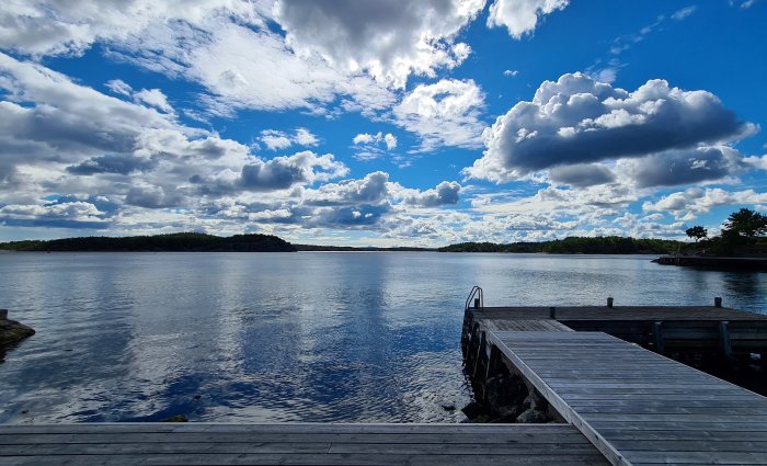 Träbrygga som leder ut till ett lugnt vatten med skogklädda öar och en dramatisk himmel med moln.