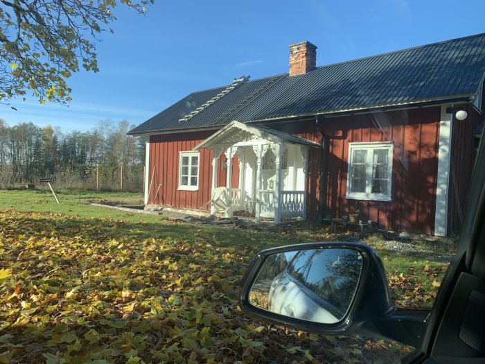 Rödmålad stuga med vit snickarglädje och löv på marken sedd från en bil.