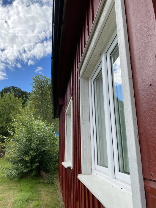 Rött trähus med vitt fönster och vindskiva, grön vegetation i bakgrunden under sommarhimlen.