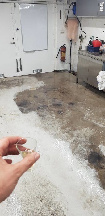 Person som håller ett glas rom i ett garage med nyligen tvättat, fläckigt betonggolv och diverse verktyg och utrustning.