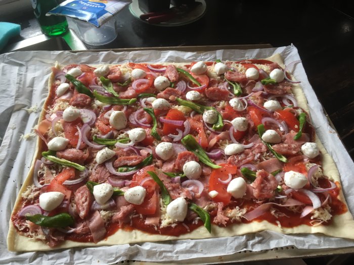 Ouppbakad pizza med tomatsås, salami, rödlök, tomat, ost, mozzarella och padrones på bakplåtspapper.