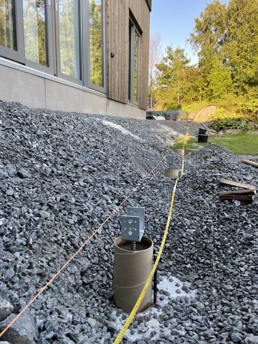 Grävda gropar med betong och stolpskor intill ett hus, redo för pergolabygge, med måttband sträckt över grus.