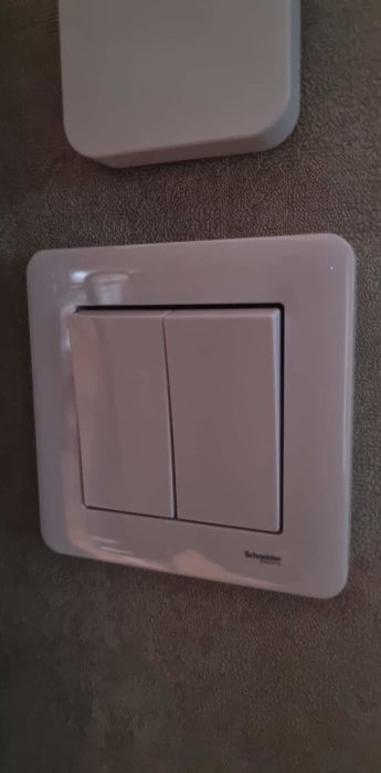 Vit dubbelströmbrytare monterad på en grå vägg, märkt Schneider.