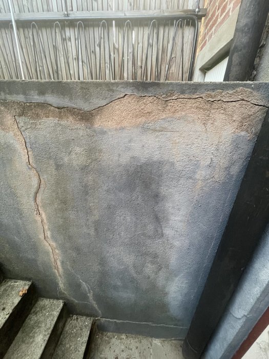 Skadad betongvägg med sprickor och avskalad färg vid trappavsats.