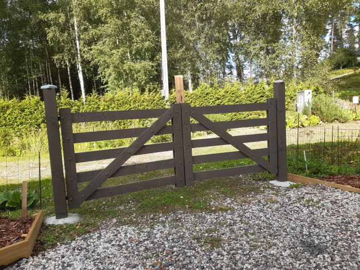 Nymålad trägrind i brunt, placerad vid en grusgång med grönska i bakgrunden.