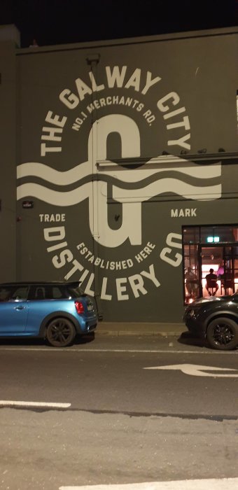 En blå bil parkerad framför en stor väggmålning med texten "The Galway City Distillery Co." på natten.