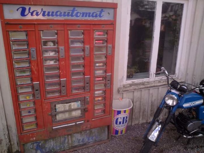 Gammal varuautomat utomhus bredvid en blå moped och en GB Glace-papperskorg.