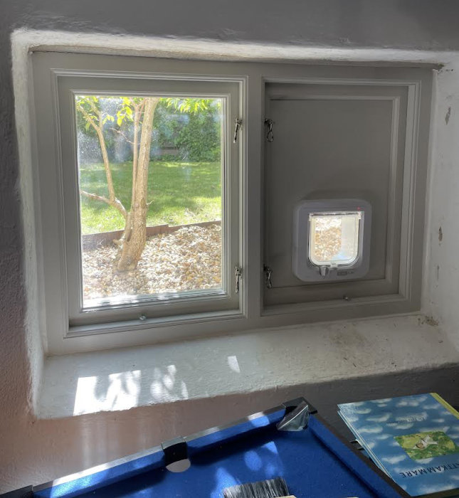 Stängt kattlucka i en gråmålad källarvägg med utsikt till en trädgård och en bok liggande på fönsterbrädan.