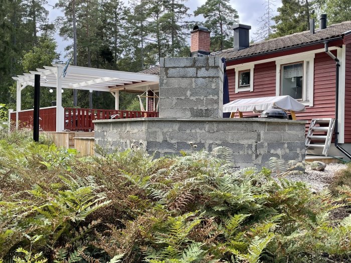 Murblock staplade på vägg av överbyggnad nära rött hus med trädäck och pergola.