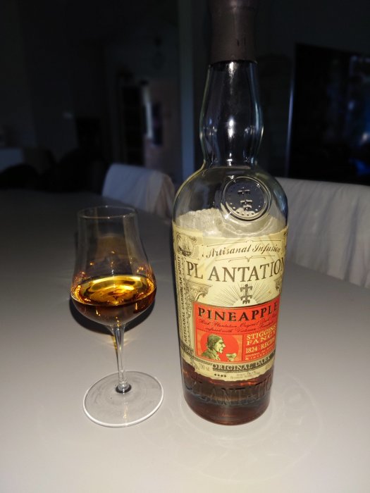 Flaska med Plantation Pineapple rom och ett glas fyllt med brun vätska framför suddig bakgrund.