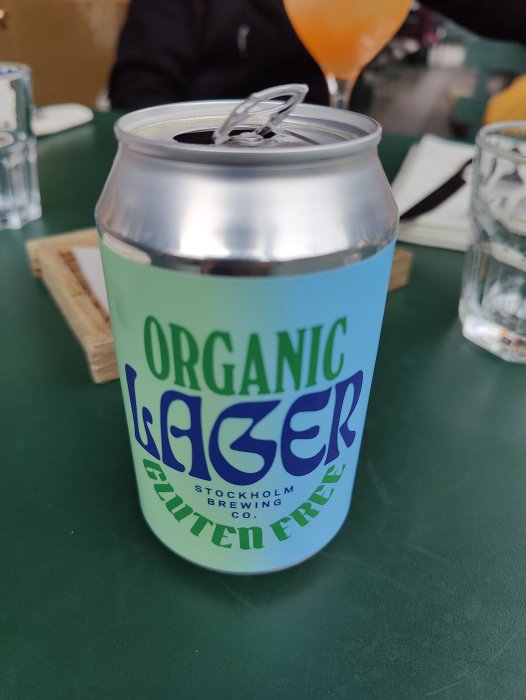 Öppnad burk av "Organic Lager" som är glutenfri på ett bord, antydan till uteservering.
