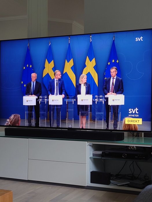 TV-skärm som visar en nyhetssändning med fyra personer stående bakom talarstolar med svenska och EU-flaggor i bakgrunden.