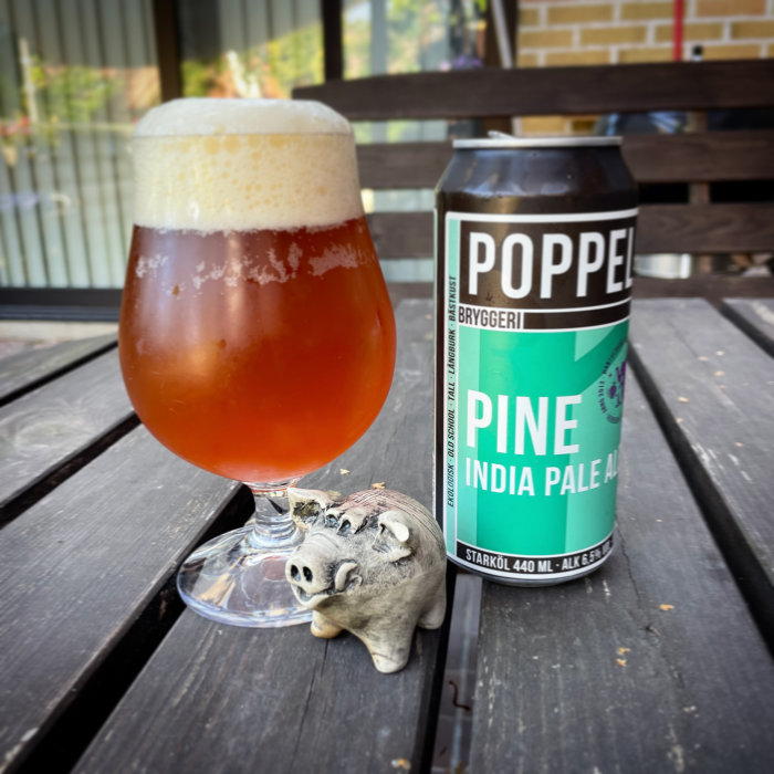 Ett glas med öl och en ölburk märkt 'Poppels Bryggeri Pine India Pale Ale' bredvid en liten grisknuten prydnad på ett träbord.