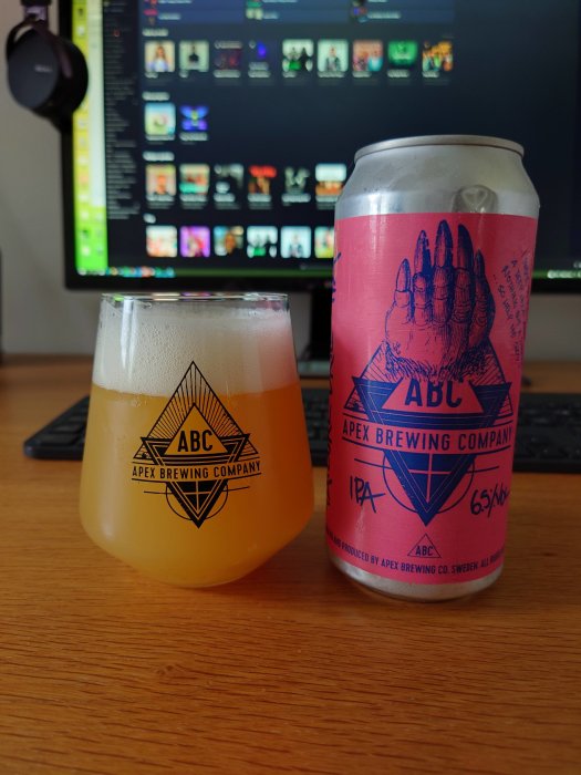 Ölglas och burk från Apex Brewing Company på skrivbord framför en datorskärm.