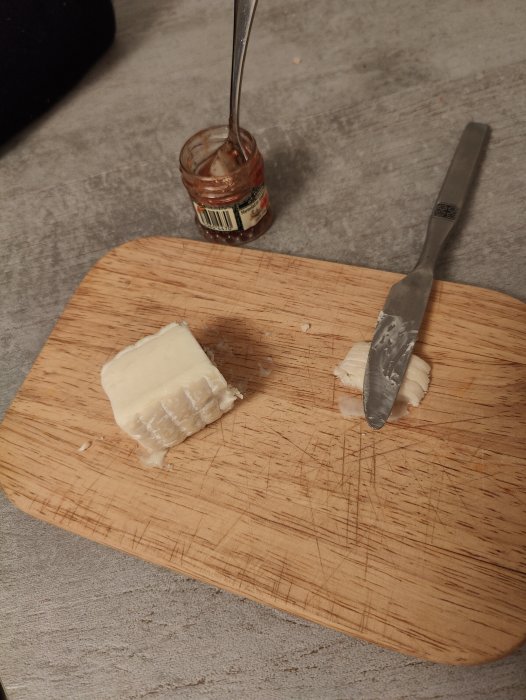 Skiva av vit ost på en skärbräda med kniv och tom marmeladburk.