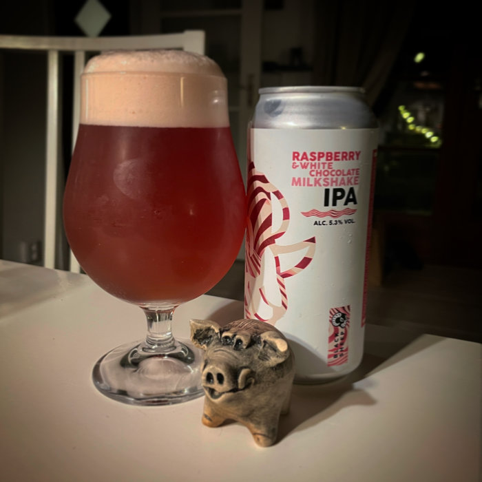 Ölglas med skum topp bredvid ölburk och en liten grisfigur på ett bord.