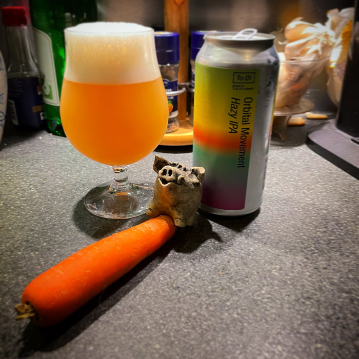 Ett glas med skummande öl, en burk IPA, och en rolig lergodsgris ovanpå en morot på köksbänk.