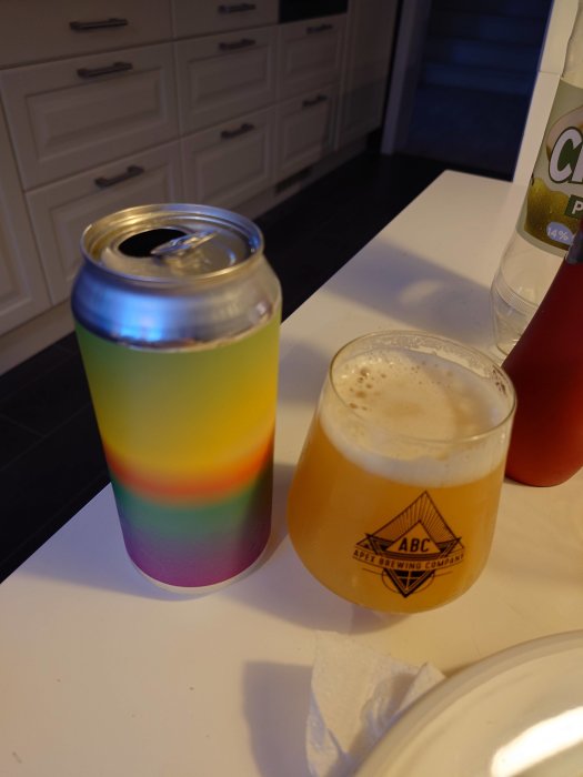 Öppnad ölburk med regnbågsmönster och en halvfull ölglas på ett kökbord.