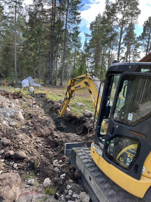 Gul minigrävare gräver utmark i skogsområde, stenar och jord syns i grävd gräv.