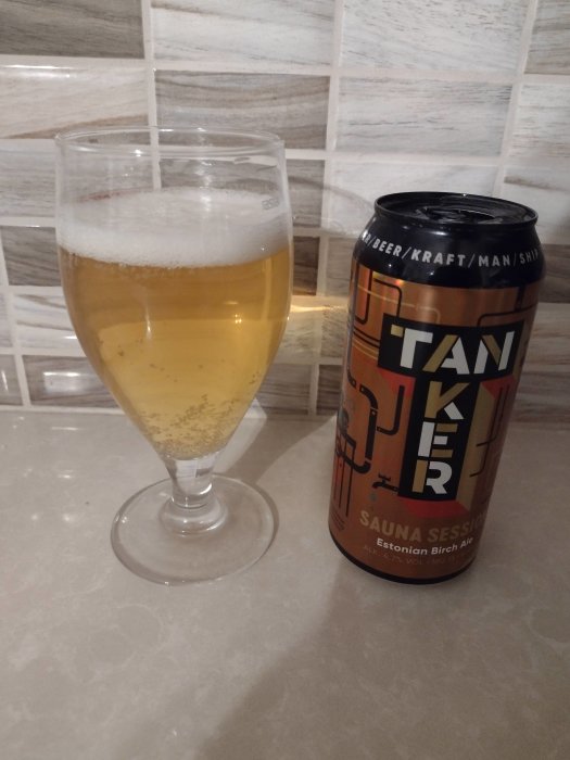 Ett glas ljus björköl bredvid en ölburk med texten "Tanker Sauna Session" mot kakelvägg.