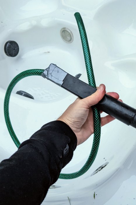 Hand håller en improviserad slang ansluten till vakuum för att suga vatten ur ett vitt SPA-bad.