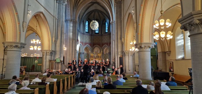 Kör och orkester framför musik i kyrkans mittgång inför publik som sitter i kyrkbänkarna.