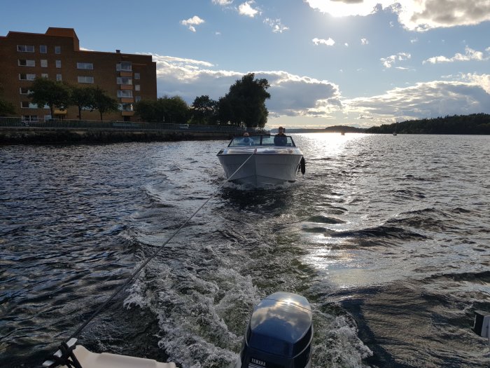 En bogserad båt på vattnet med en annan båt och strand i bakgrunden.