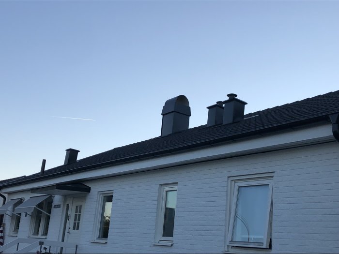 Ett vitputsat hus med svart tak och skorstenar mot en klar himmel vid skymningen.