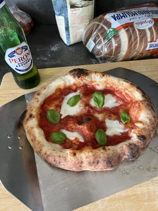 Hemlagad pizza med tomat, mozzarella och basilika på spade, bredvid Peroni-öl och brödpaket.